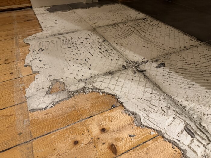 Gammalt furugolv med rester av bruk och kakel ovanpå. Delvis borttagna klinkerplattor visar nät och bruk kvar på golvytan.