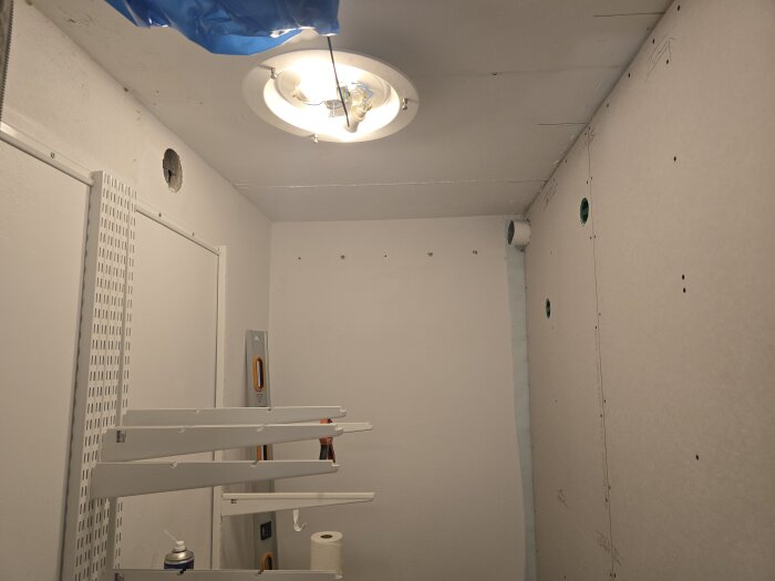 Klädkammare med vitmålade väggar och tak, ventilation och spirorör installerade. Hål i väggar för röranslutningar. Ljusramp i taket. Hyllor och verktyg syns.