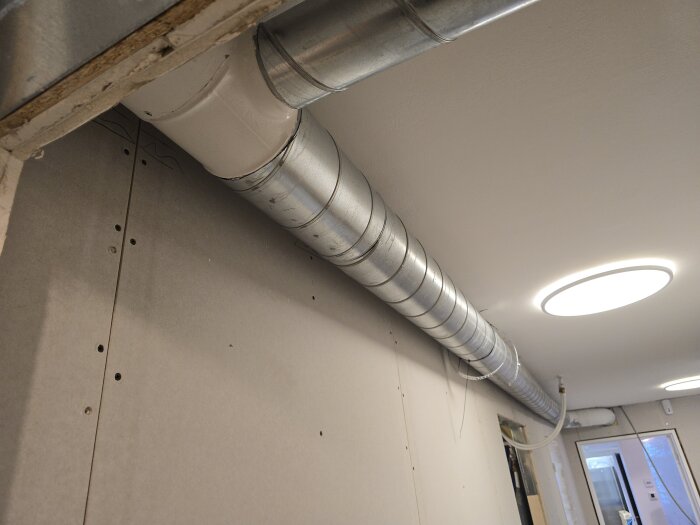 Ventilationsrör monterade längs taket i en korridor med vita takspotlights. Väggen är delvis täckt med oskivade gipsskivor.
