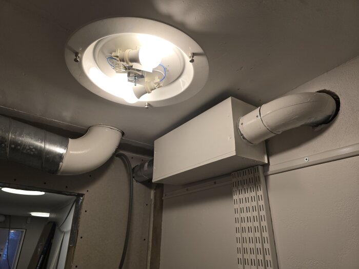 Ventilationsrör och installation i ett rum med synliga spirorör och lampa nära taket. Rören går genom väggen och är delar av ett renoveringsprojekt.