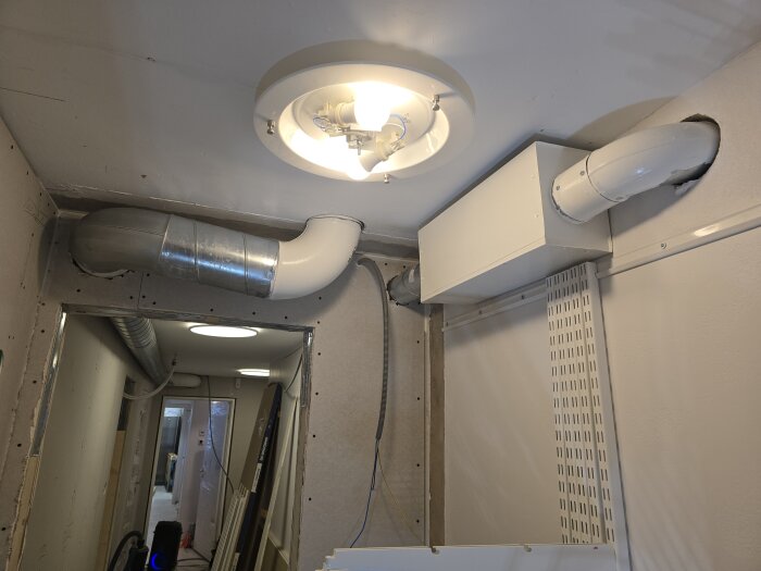 Klädkammare med synliga ventilationsrör och ljusarmatur i taket, ny ventilation installerad genom väggarna.