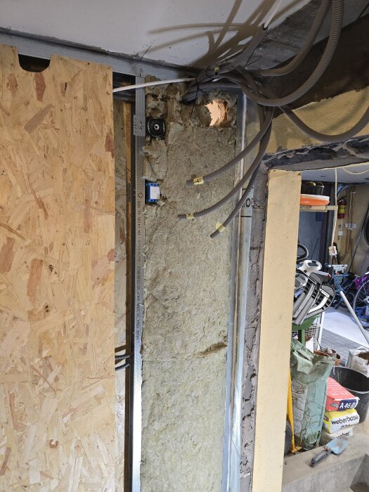 Byggkonstruktion med exponerad isolering och flexrör, delvis täckt med OSB-skiva. Synliga verktyg och byggmaterial på golvet i bakgrunden.