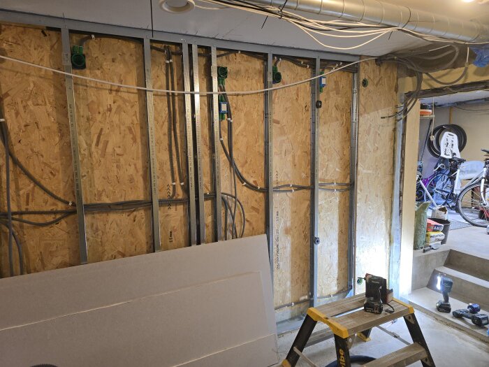 Vägg med synliga flexrör för ventilation och CAT6-kablar bredvid en stege; OSB-skivor och verktyg synliga i ett pågående byggprojekt.