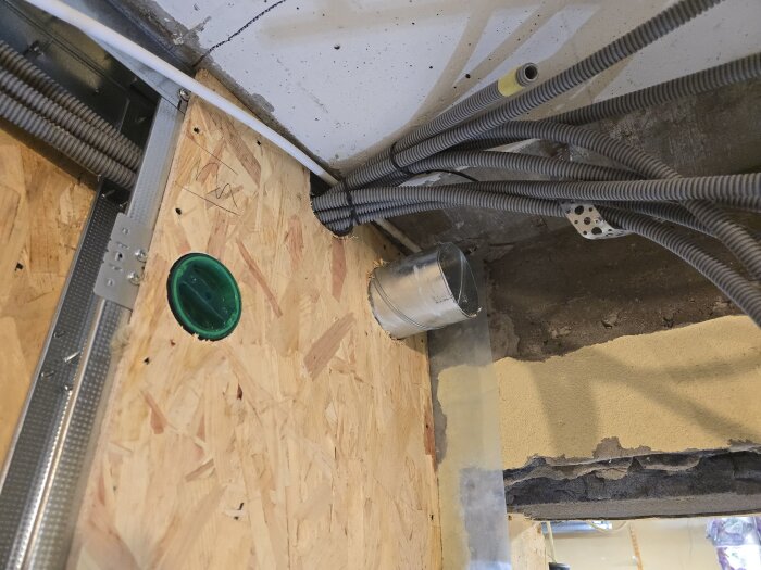 Ventilationsrör och flexrör monterade i en öppning med träpanel och betongvägg, med synliga skruvclips och ett grönt eluttag.