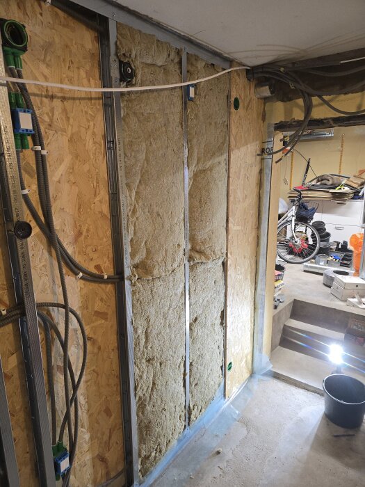 Vägg med isolering, flexrör för CAT6-kablar och byggmaterial i en renoveringsmiljö, stege och cykel synliga i bakgrunden.