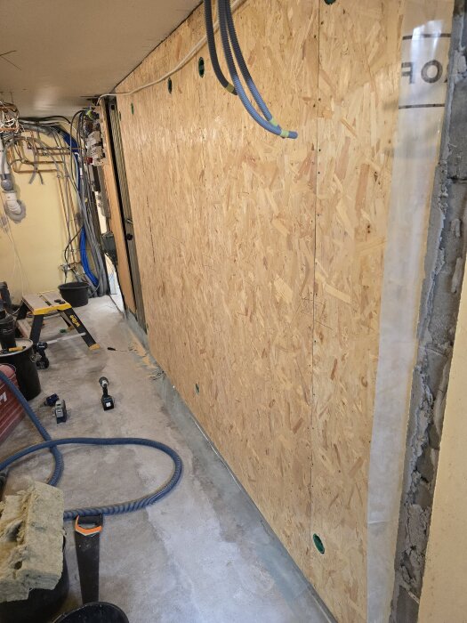 Vägg i källare med ny OSB-skiva och synliga flexrör för CAT6-kablar, verktyg och byggmaterial på golvet. Montering och bygge pågår.
