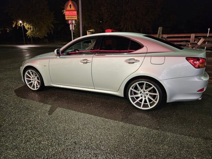 Silverfärgad bil parkerad på våt asfalt under natten, delvis belyst av en gatulampa, med trafikskyltar i bakgrunden.