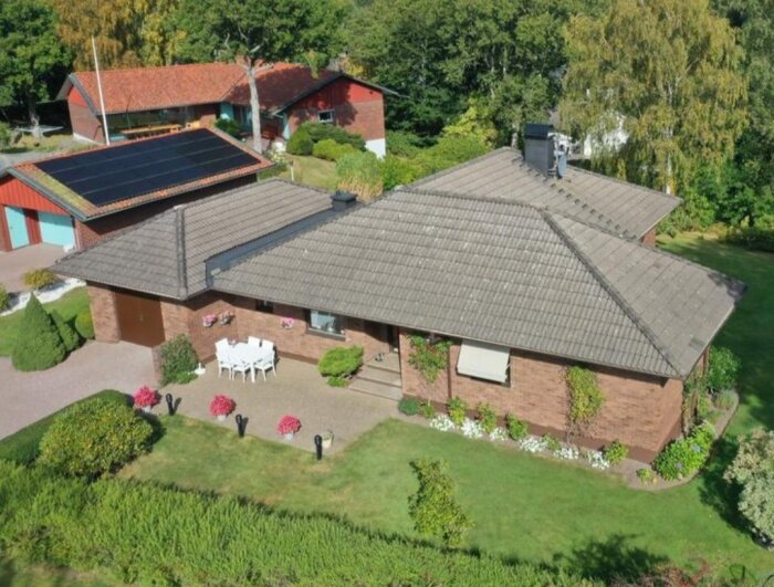 Flygbild på ett hus med nyant målat mörkt tak, omgiven av välskött trädgård med buskar och blommor, en uteplats med möbler synlig.