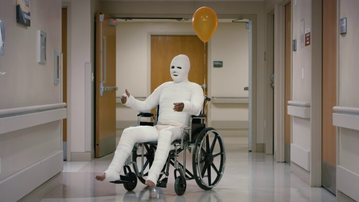 En person helt täckt i bandage sitter i rullstol med en orange ballong i en sjukhuskorridor.