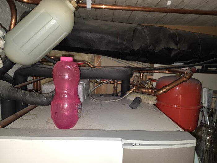Översiktsbild av värmesystem med rör, röd expansionskärl och ett svart isolerat rör ovanpå en värmepump, en rosa flaska i förgrunden.