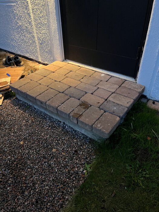 Gamla stenplattor vid en ytterdörr, ovanpå en betongplatta, omgiven av grus och gräs i skymningsljus.