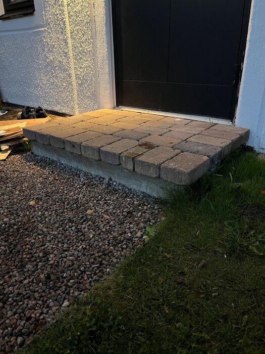 Gamla stentrappsteg framför en dörr, ovanpå en betongplatta, omgivna av grus och gräs.