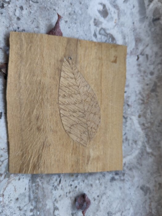 Intarsia på träplatta med bladmotiv ligger på stenunderlag.