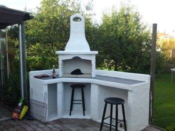 Utomhusgrill i vitt stenmaterial med skorsten, omgiven av två svarta stolar på en stenbelagd uteplats, omgiven av grönska.