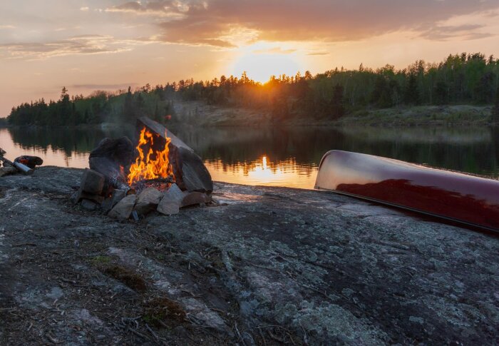 Lägereld vid sjö vid solnedgång, omgiven av stenar och en uppochnedvänd kanot på klippa.