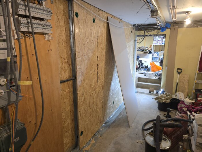 Byggarbetsplats med OSB-skivor, gipsskiva lutad mot väggen, ventilationsrör i taket och verktyg på golvet. Elektrisk panel synlig på väggen.