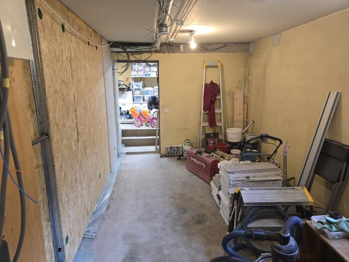 Källarutrymme under renovering, med verktyg, byggmaterial och stege. OSB-vägg, målade väggar och en leksakssparkbil i bakgrunden.