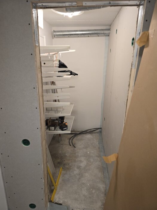 Renovering av klädkammare, oisolerade väggar med hyllor, ventilation rör, verktyg och material på golvet, belysning och betonggolv syns.