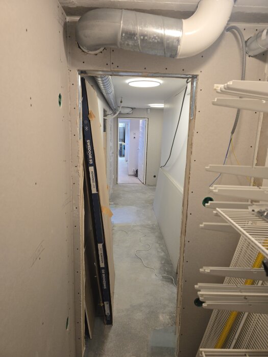 Renoverad korridor med gipsskivor, synliga ventilationsrör och eldragning. Dörröppning leder till ett upplyst rum i bakgrunden. Byggmaterial lutar mot väggen.