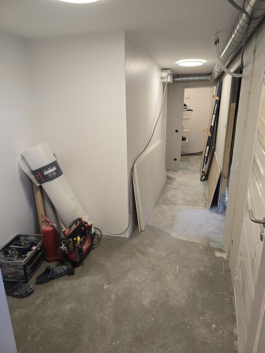 Renovering av korridor med gipsskivor, verktygslåda och brandsläckare synliga, ventilationsrör längs taket. Snickarverktyg och kabel på golvet.