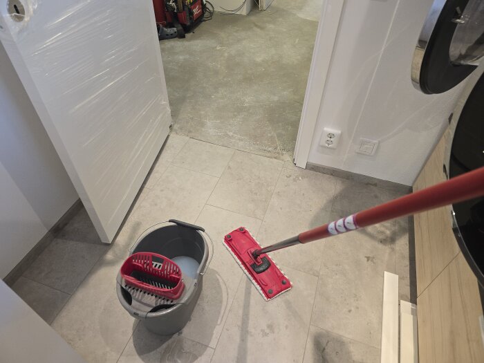 En mopp och hink står i ett rum med kaklat golv. En dörr står uppställd mot en vägg, och byggmaterial syns i bakgrunden.