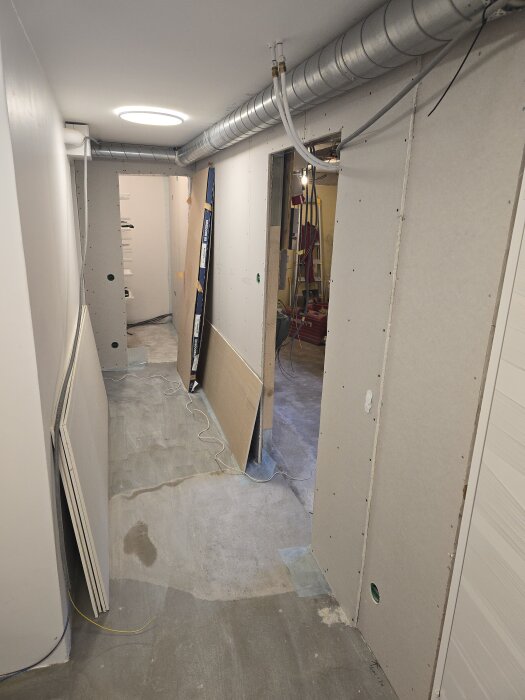 Renoverad korridor med nyligen installerade gipsskivor och synliga spirorör för ventilation. Byggmaterial och verktyg står längs väggarna.