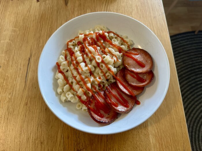 Tallrik med makaroner och skivad korv toppade med ketchup, serverad på ett träbord.