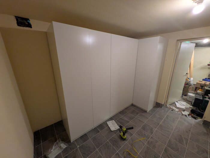 Tvättstuga med IKEA PAX-garderob, hörn med ventil i taket, verktyg och papper på golvet, dörröppning till rum med kartonger.