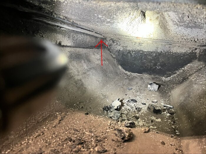 Inuti en vedpanna syns en yta med smuts och en röd pil som pekar på en spricka där det läcker, omgiven av sot och aska.