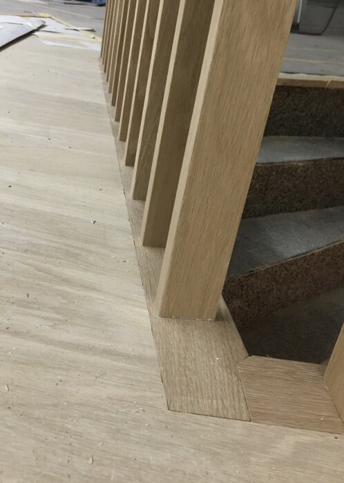 Träspaljé under konstruktion, visar vertikala trästolpar infällda i ett ljust trägolv, placerade vid en trappa.