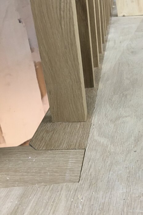 Närbild på en träspaljé med vertikala träpinnar som monterats i en golvsockel av samma material. Golvet ser ut att vara av trä.