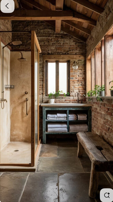 Rustikt badrum med tumlat stengolv, dusch med glasväggar, och trädetaljer samt en bänk och handdukar på hylla under fönster.