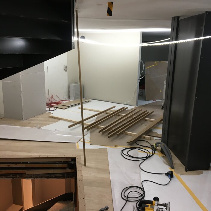 Rum under konstruktion med träplankor på golvet, en stav upprätt, svart trappa till vänster och byggutrustning runtomkring.