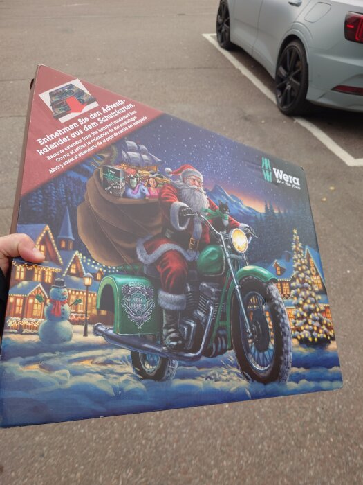 Adventskalender med tomte på motorcykel, julmotiv med snögubbe och dekorerade hus, hålls i handen nära en parkerad bil.