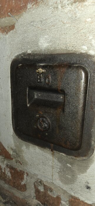 Gjutjärnslucka med en skruv fastsatt i ett murat väggparti, delvis rostig yta.