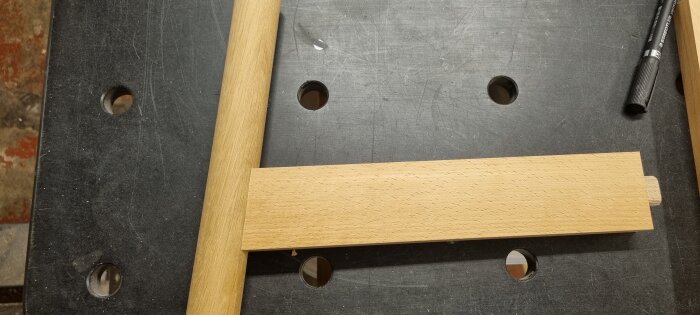 En bit trä med en rund stav visas på en arbetsbänk. En penna ligger på bänken.