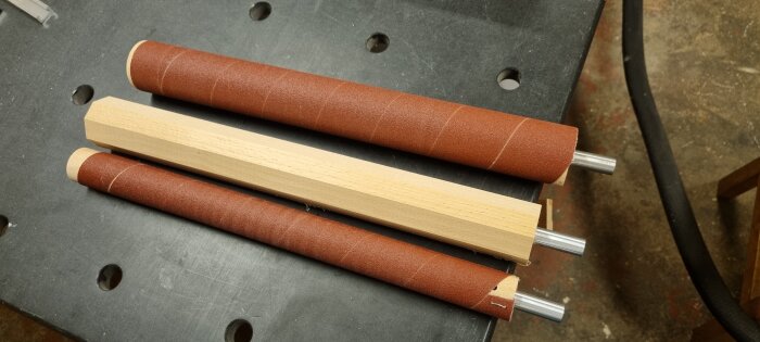 Sliprullar i varierande storlek, några täckta med sandpapper och en åttkantig trästav, ligger på en arbetsbänk med metalldetaljer synliga.