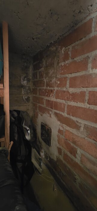 Röd tegelvägg med igenmurat hål ovanför sotlucka; hylla med prylar syns delvis.