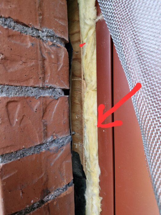 Tegelfasad med synlig isolering och plåt vid dörrkarm, röd pil markerar området där plåten ansluter mellan tegel och isolering.