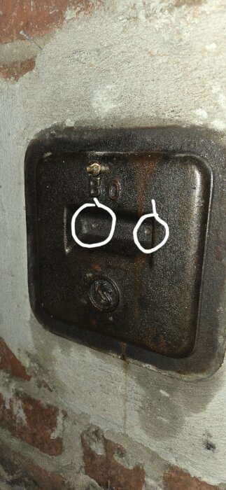 Närbild av en svart, kvadratisk lucka av metall med två täckta skruvar överst och vita markeringar runt skruvarna, omgiven av betong och tegel.