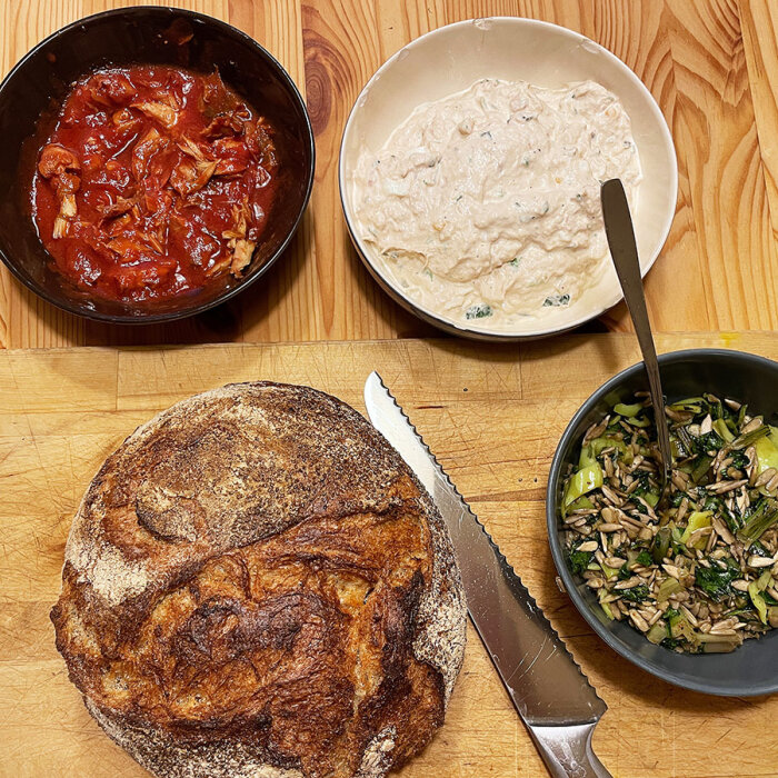 Bröd med en kniv på skärbräda. Skålar med makrill i tomatsås, tonfiskröra och grönsaksblandning på ett träbord.