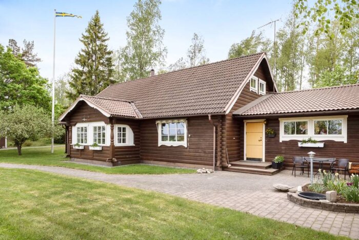 Brunt timmerhus på landet med vita detaljer och brunt tak, omgiven av grönska, med trädgårdsmöbler och svensk flagga i bakgrunden.