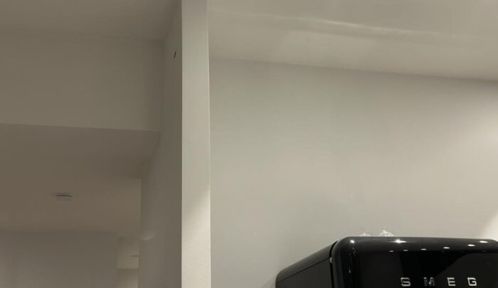 Vy från köket mot hallen, visar en vit vägg och toppen av ett svart kylskåp märkt "Smeg".