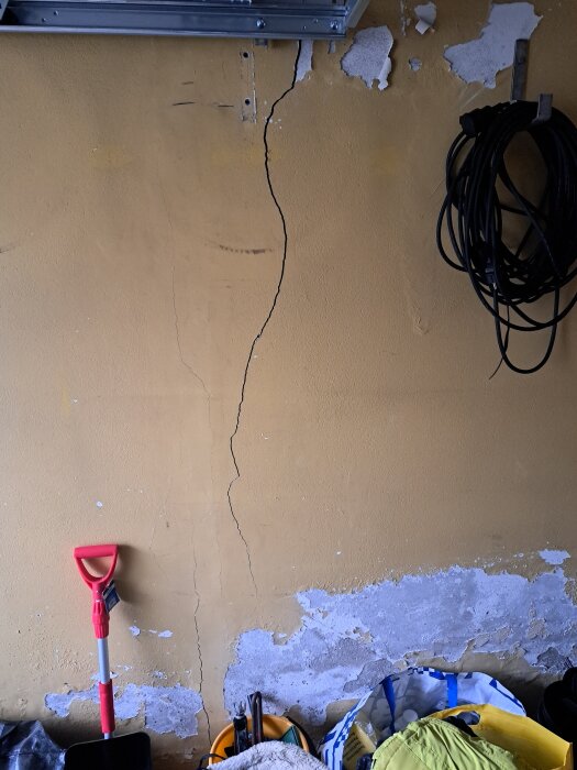 Spricka i en gul garagevägg med avflagad färg. Till höger syns ihoprullad kabel på väggen och till vänster står en spade.