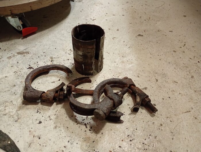 Rostiga avgasrörsklammer och ett kort, rostigt rör ligger på ett golv, del av ett avgasrörsprojekt för en Volvo.