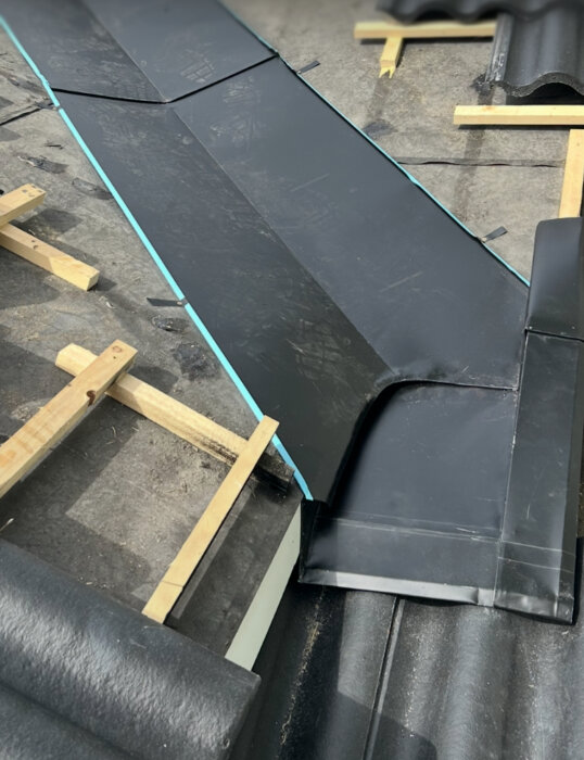 Svart plåt uppvikt runt ränndalsplåt på tak, delvis synligt bärläkt, ej slutläktat, med träläkt som sticker ut över ränndalsplåten.