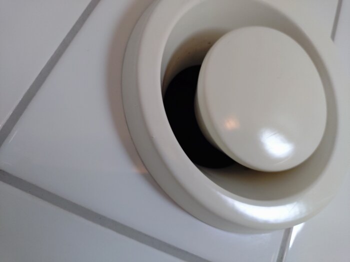 Vit ventilationstrumma med lös, snedställd kon i mitten, monterad på vit kaklad vägg.