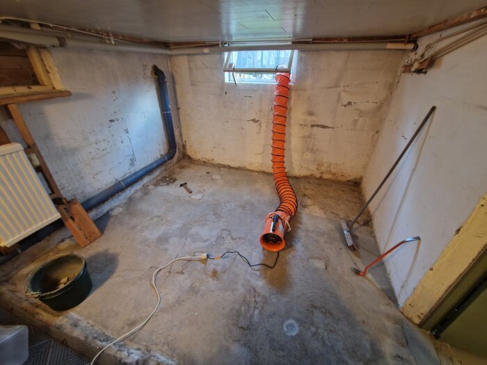 Källarrum med bilat betonggolv, orange ventilationsslang och synliga rör längs väggen; verktyg och en skopa på golvet.