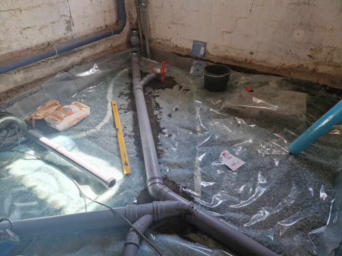 VVS-installation i källare, grus och rörsystem på plastfolie, avloppsrör, vattenpass, cementpåse och hink på betonggolv under renovering.