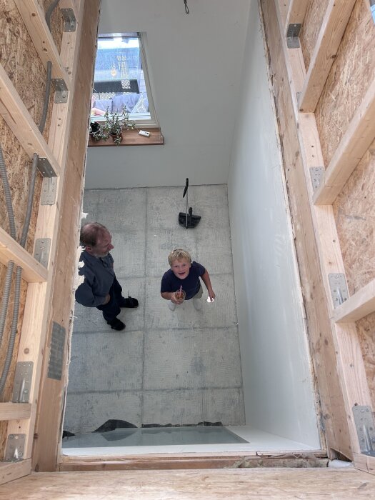 Två personer tittar upp genom ett hål i taket, som är upptaget för en trappa. Enheten har vitmålad vägg, träreglar och ett fönster med växter.